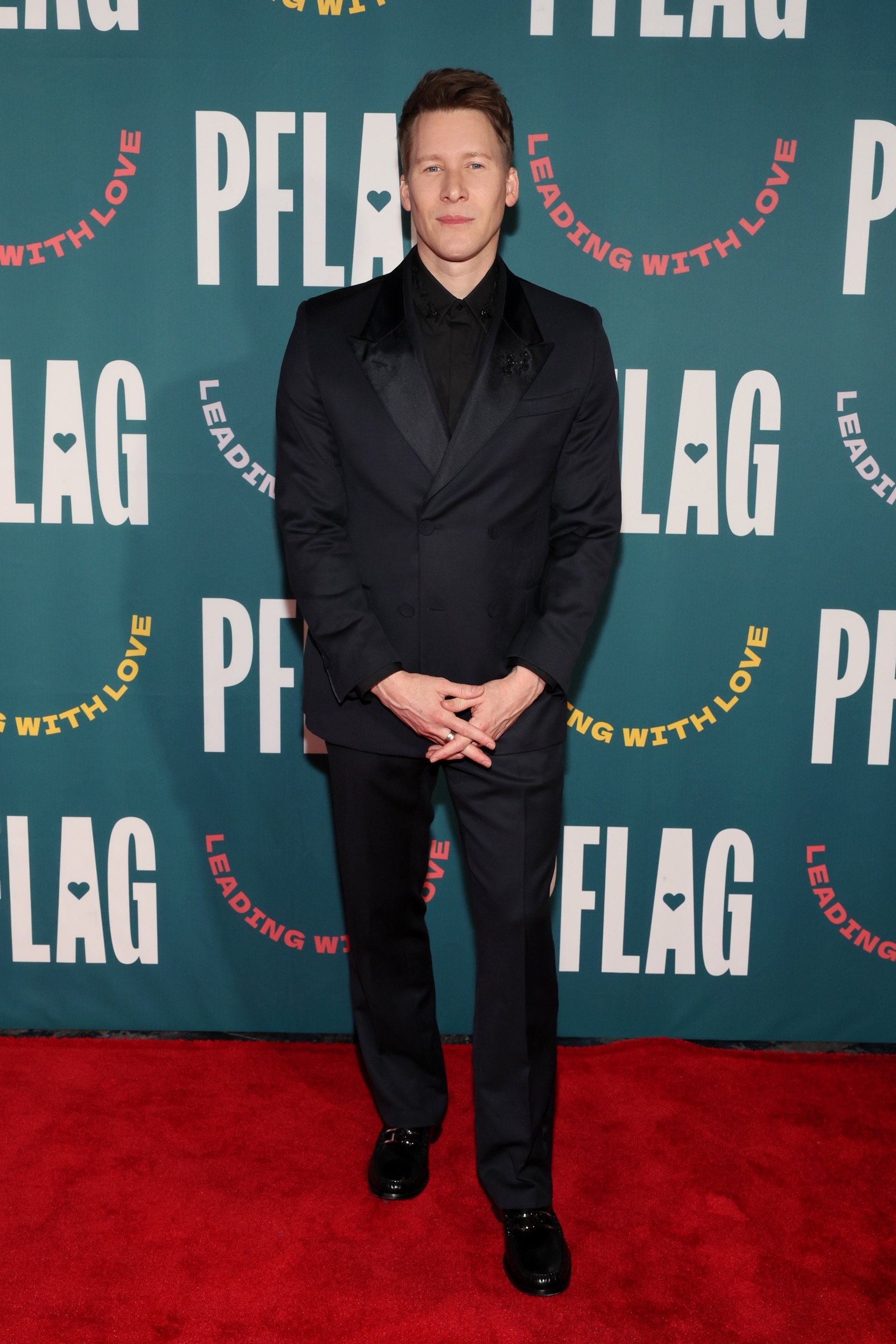 Adult man in a dark suit on a red carpet in front of a PFLAG banner. He is looking forward with his hands clasped.