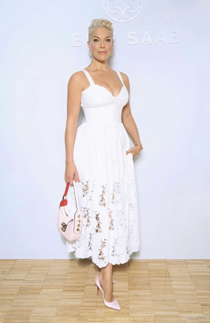 Elegant woman in a white dress with floral embroidery, accessorized with a light pink handbag and matching heels, poses against a white backdrop.