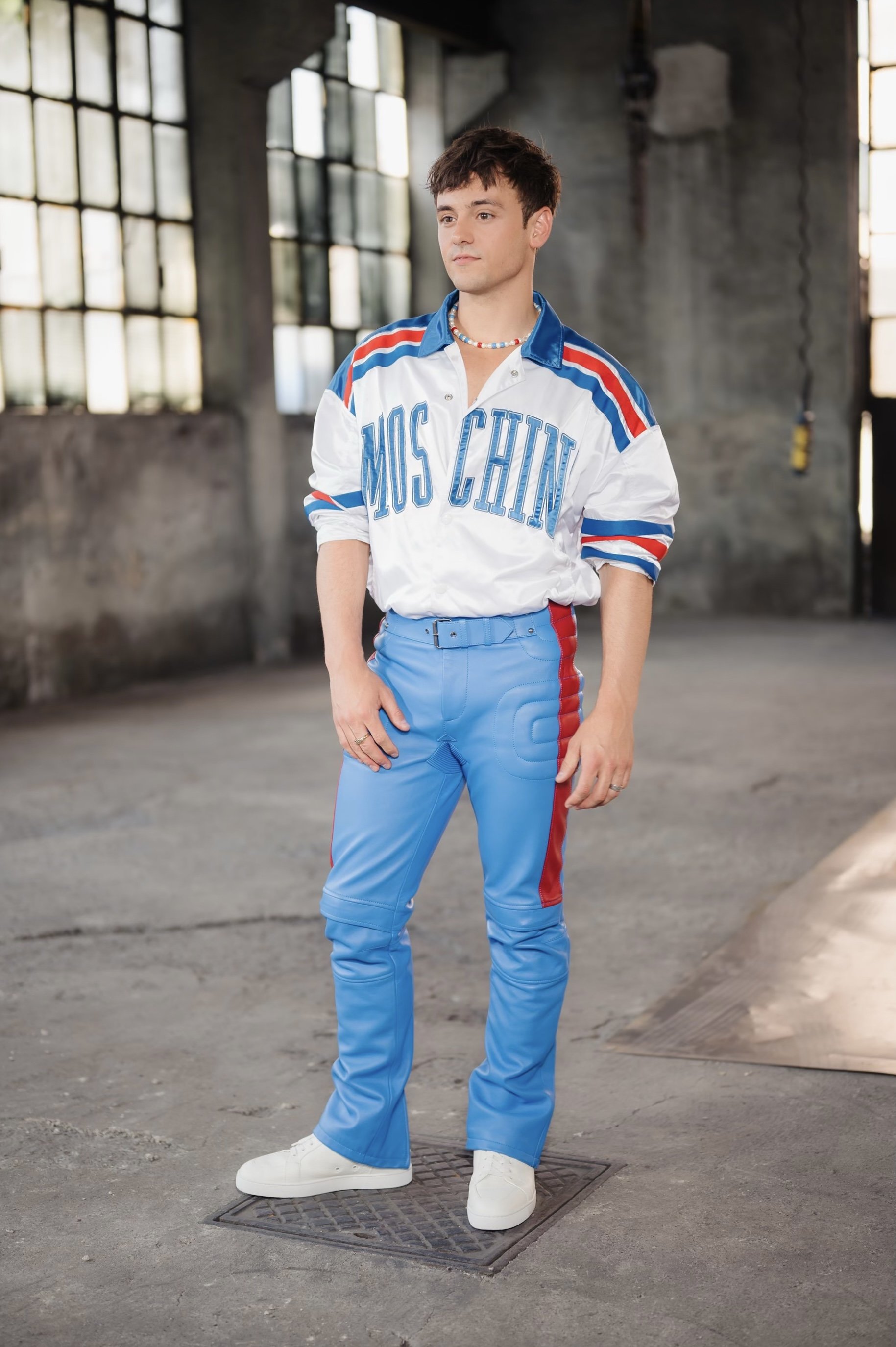 Young adult male in a Moschino shirt and blue pants poses in a gritty urban interior with large windows and textured concrete walls.