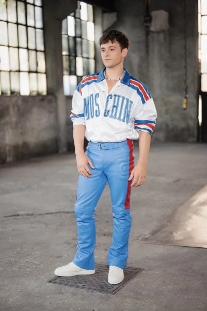 Young adult male in a Moschino shirt and blue pants poses in a gritty urban interior with large windows and textured concrete walls.