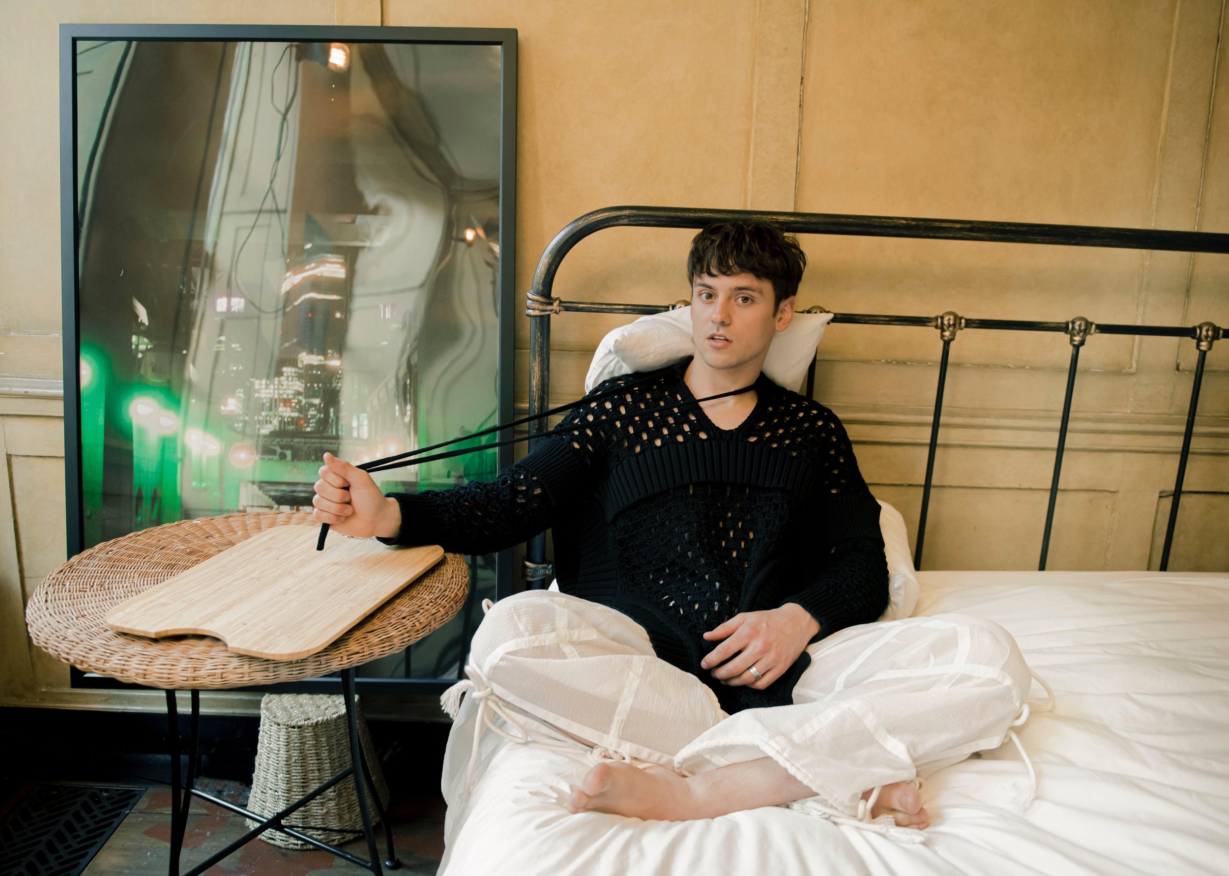 Young man poses on a bed with an abstract painting, wearing a black knit sweater and white pants in a cozy indoor setting.