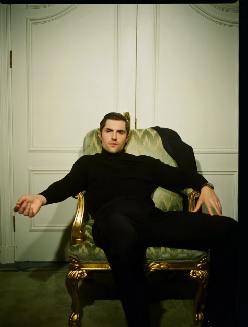 Elegant man in a black turtleneck and pants sits in a gold-trimmed, green armchair against a white door backdrop in a portrait-style setting.