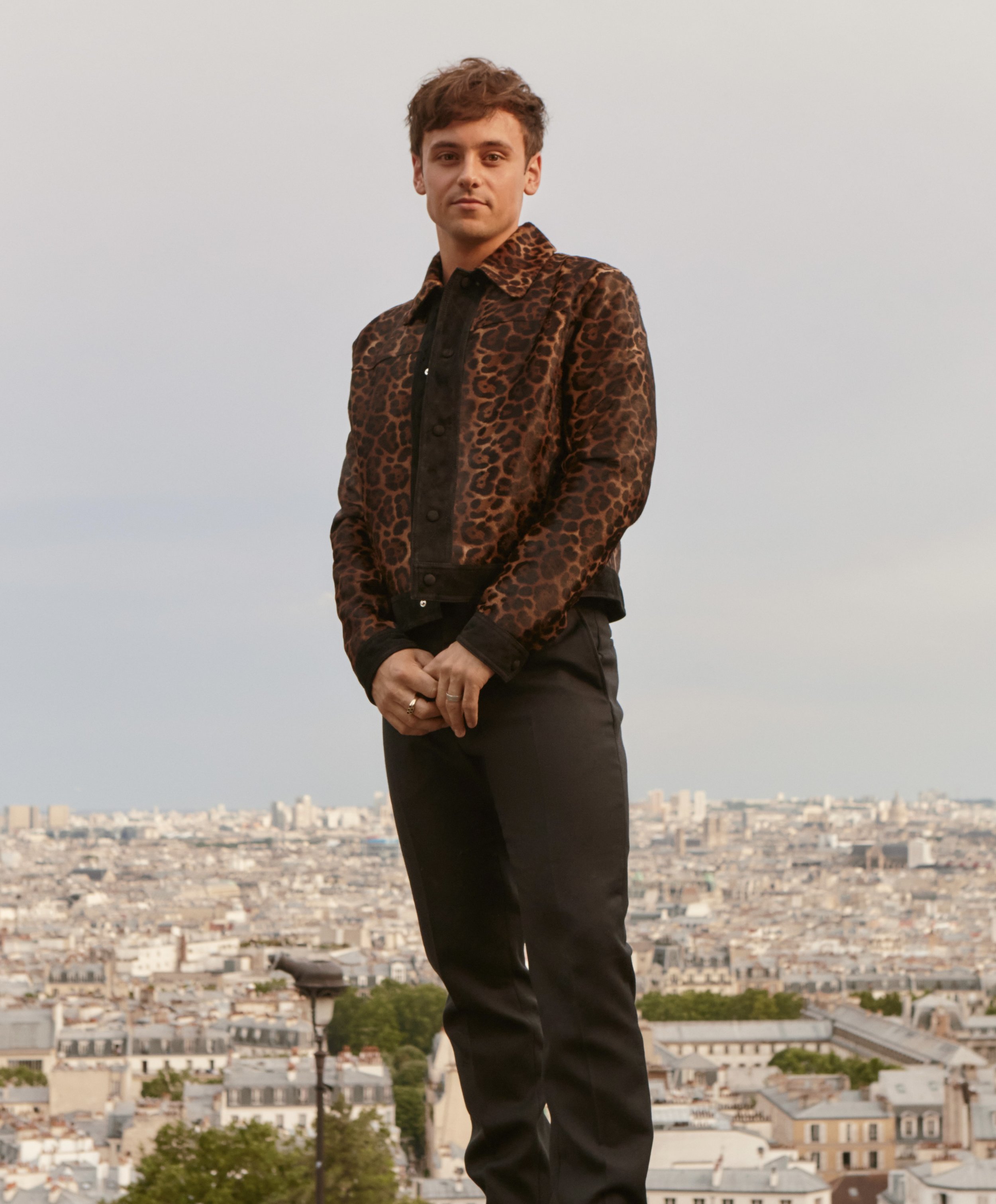Portrait of a man standing outdoors against a cityscape backdrop. He wears a brown leopard-print shirt with black pants.