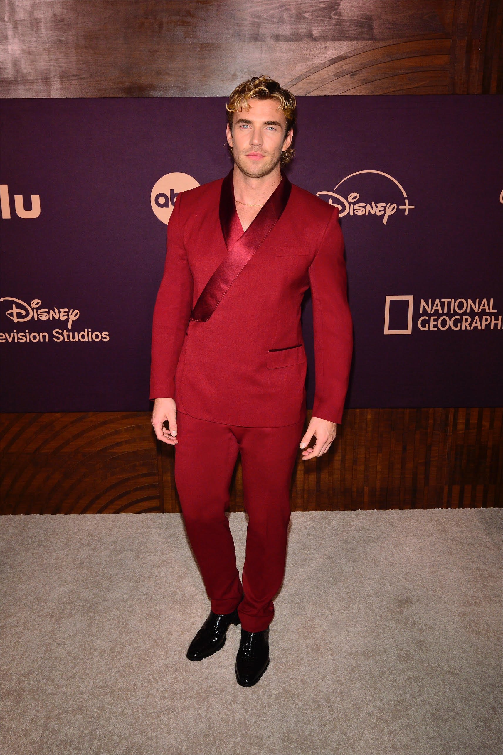 Full-length shot of a man in a red suit standing in front of a branded event backdrop. He has blond hair and black dress shoes.