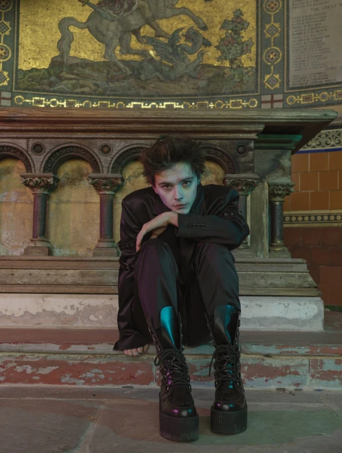 Young person in platform boots sits on stone steps indoors with an ornate background in a dimly lit setting with diffused light.