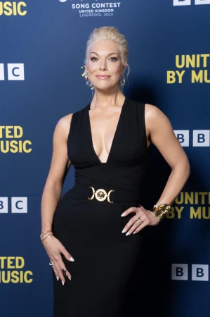 Blonde woman in elegant black dress on navy backdrop promoting Eurovision Song Contest in Liverpool, United Kingdom. Formal wear, jewelry.