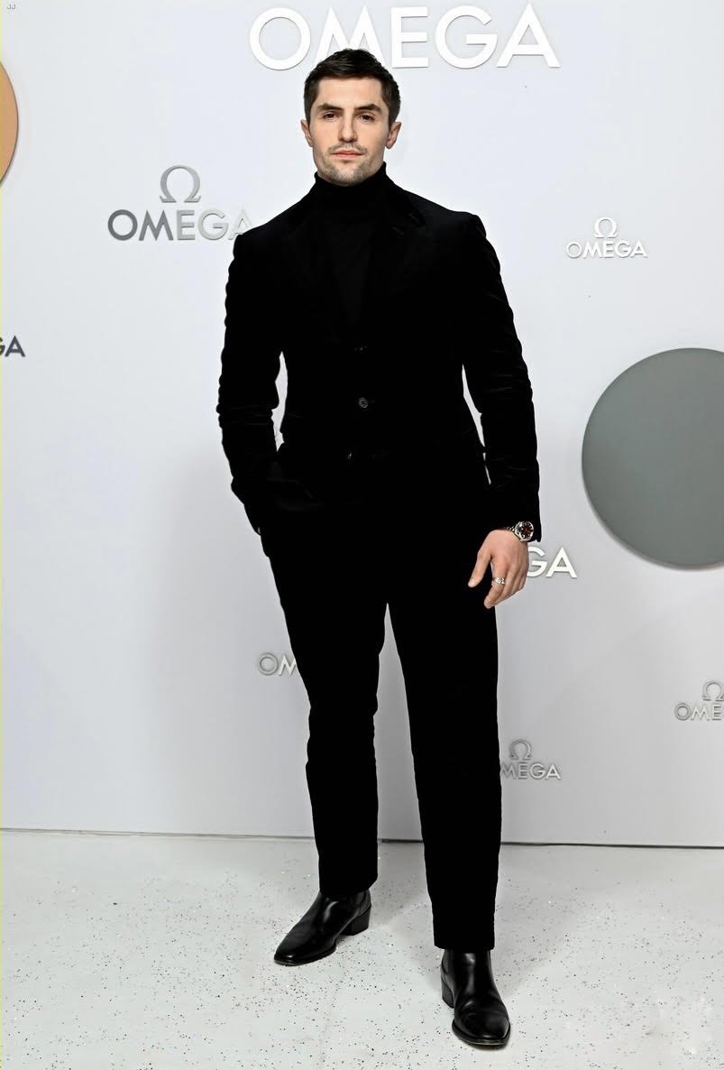 A man in a black suit stands against a white backdrop featuring the Omega logo. He poses in a full shot, wearing a black turtleneck.