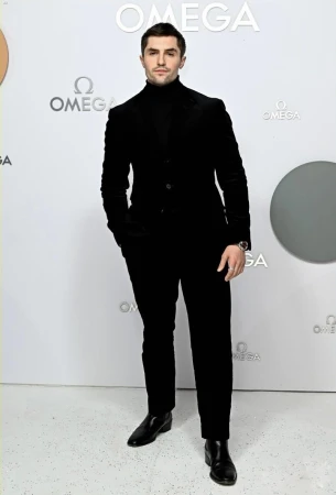 A man in a black suit stands against a white backdrop featuring the Omega logo. He poses in a full shot, wearing a black turtleneck.