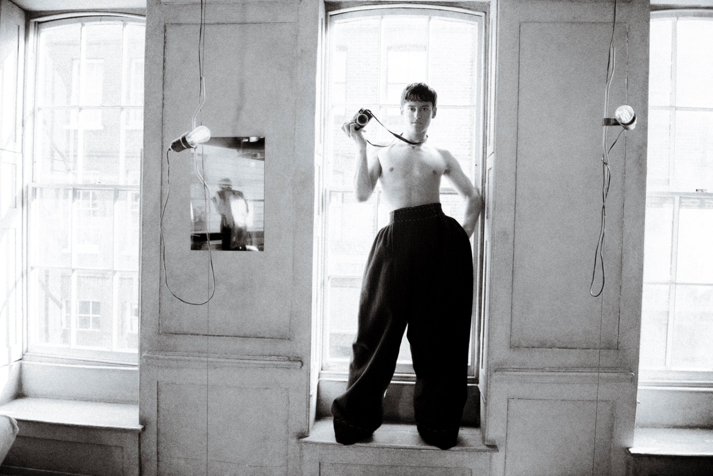 Monochrome portrait of a young adult standing topless in window with digital camera, large pants, and bare bulb hanging from the ceiling.