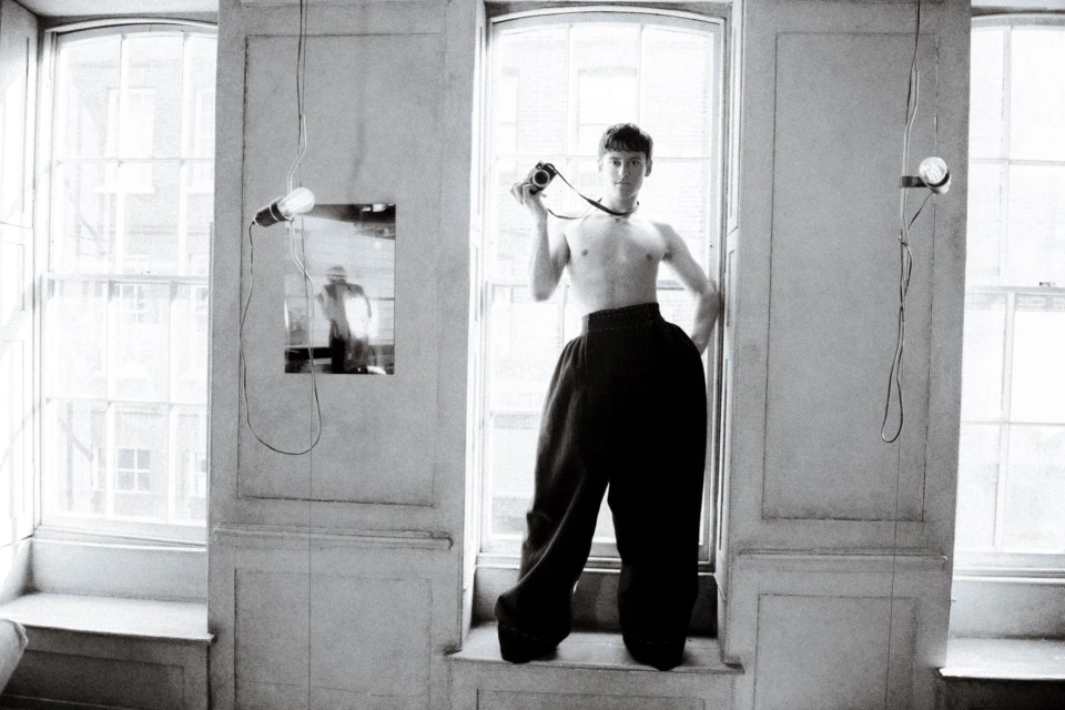 Monochrome portrait of a young adult standing topless in window with digital camera, large pants, and bare bulb hanging from the ceiling.