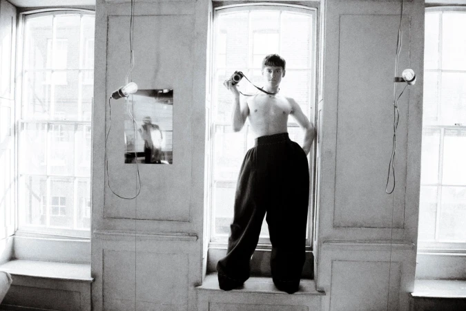 Monochrome portrait of a young adult standing topless in window with digital camera, large pants, and bare bulb hanging from the ceiling.