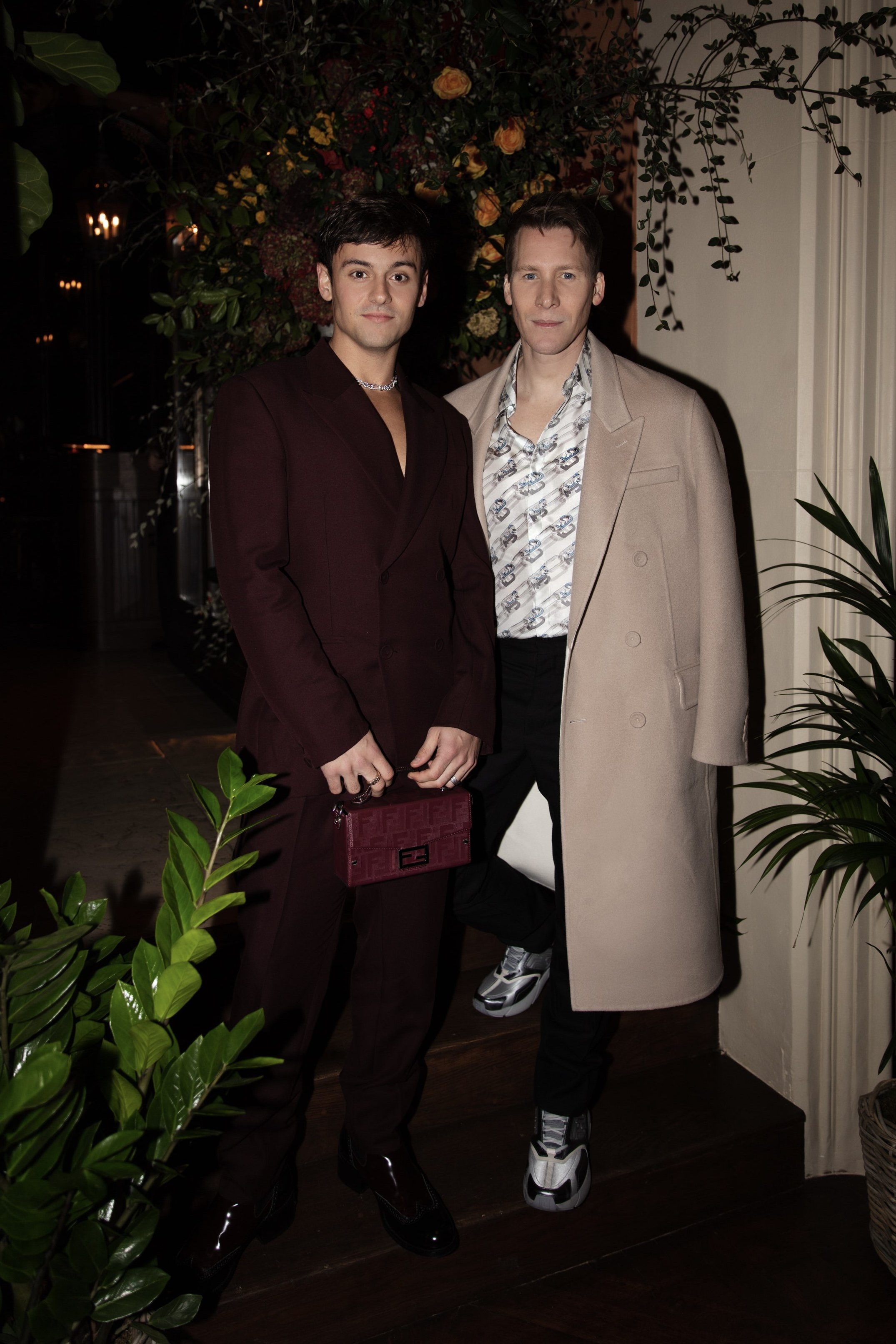 Two men pose in formal attire at an indoor event with floral decorations and plants, one in a suit with a bag and the other in a coat.