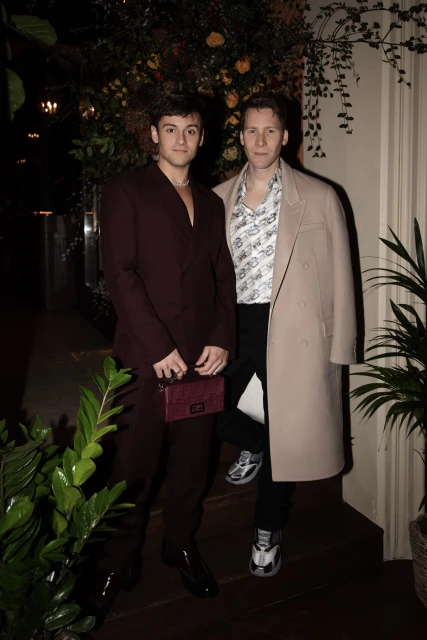 Two men pose in formal attire at an indoor event with floral decorations and plants, one in a suit with a bag and the other in a coat.