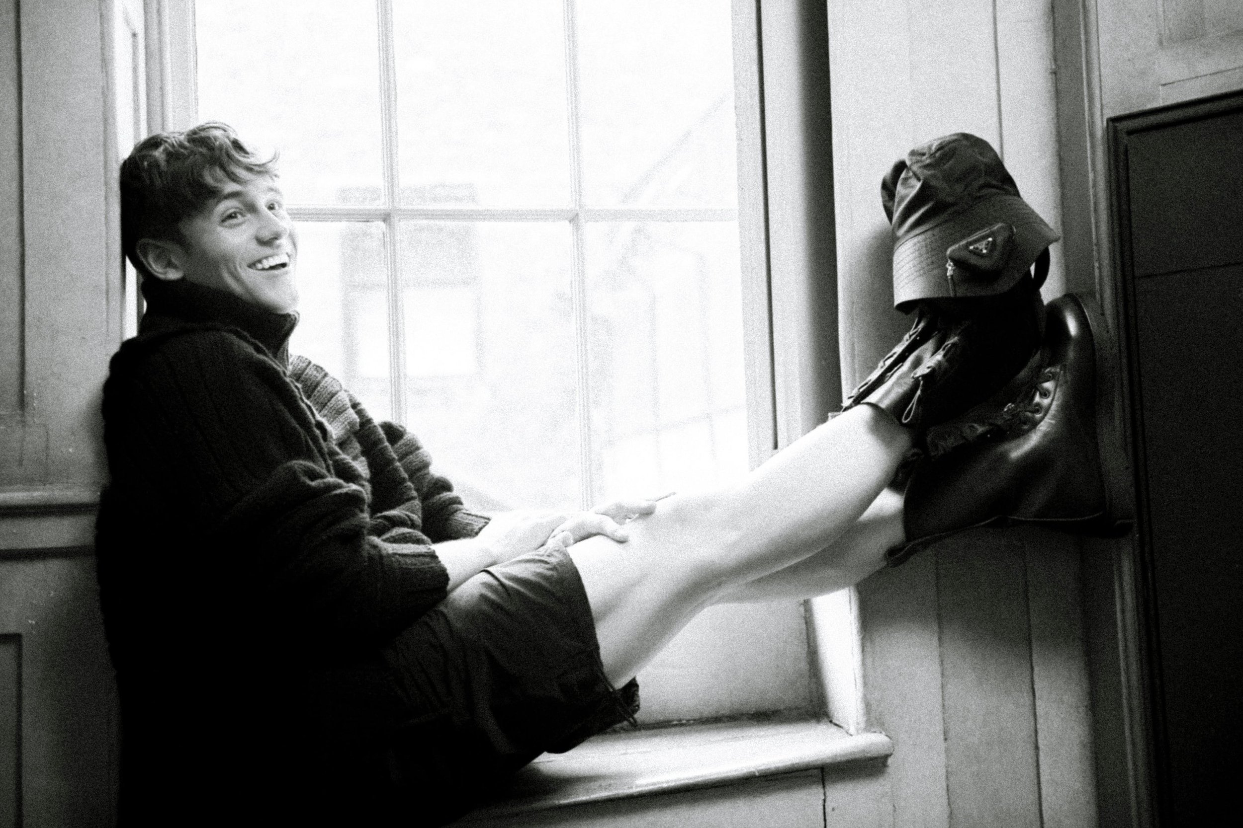 Smiling young man in a dark turtleneck sweater seated on a deep window sill with his legs propped up against the wall