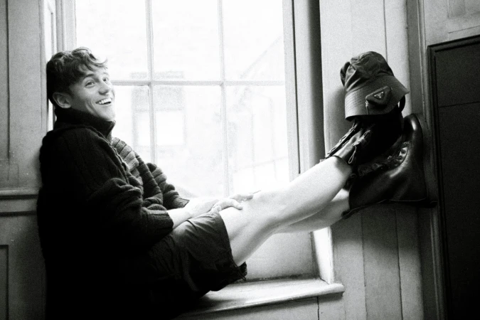 Smiling young man in a dark turtleneck sweater seated on a deep window sill with his legs propped up against the wall
