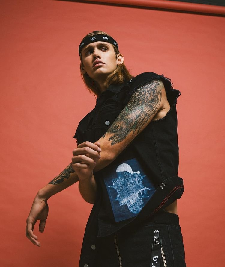 Portrait of a blonde male model with a bandana wearing a black vest. The backdrop is a solid coral color, with soft, diffused lighting.