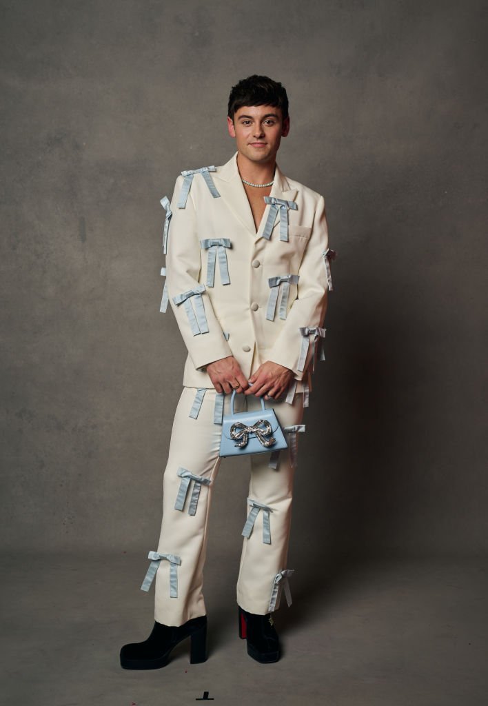 Portrait of a young adult male in a custom cream suit adorned with light blue bows, holding a matching purse, and wearing black platform boots.