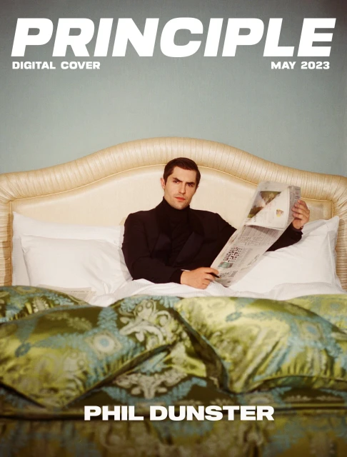 Magazine cover features a man in a black suit reading a newspaper in bed with a decorative headboard and patterned bedding, 2023.