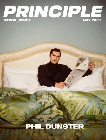 Magazine cover features a man in a black suit reading a newspaper in bed with a decorative headboard and patterned bedding, 2023.