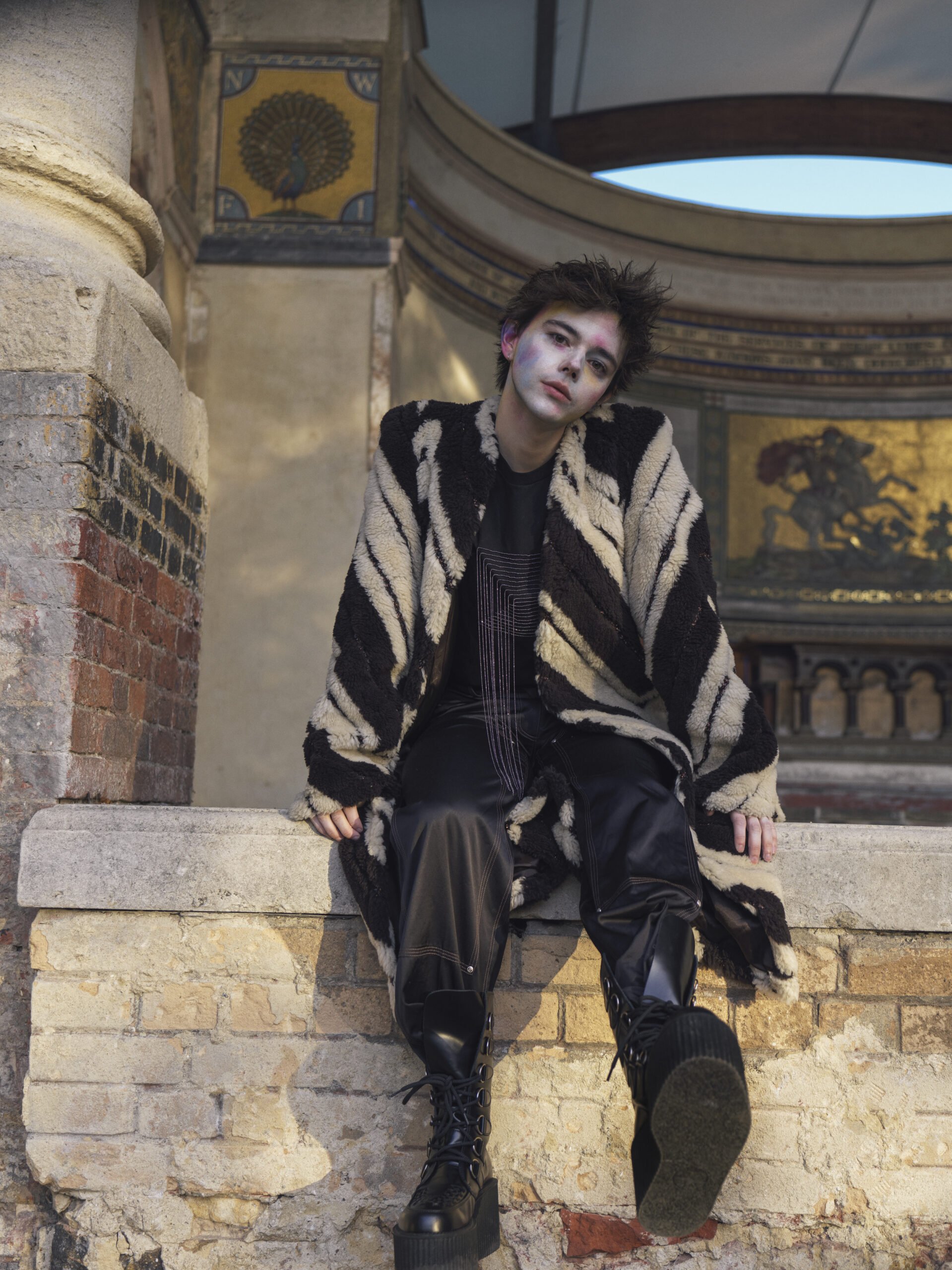 A person with makeup sits outside, wearing an animal-print coat and boots, with a brick wall, architectural details, and sky in background.