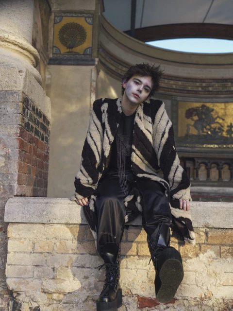 A person with makeup sits outside, wearing an animal-print coat and boots, with a brick wall, architectural details, and sky in background.