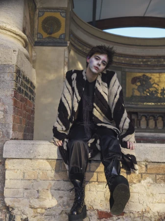 A person with makeup sits outside, wearing an animal-print coat and boots, with a brick wall, architectural details, and sky in background.
