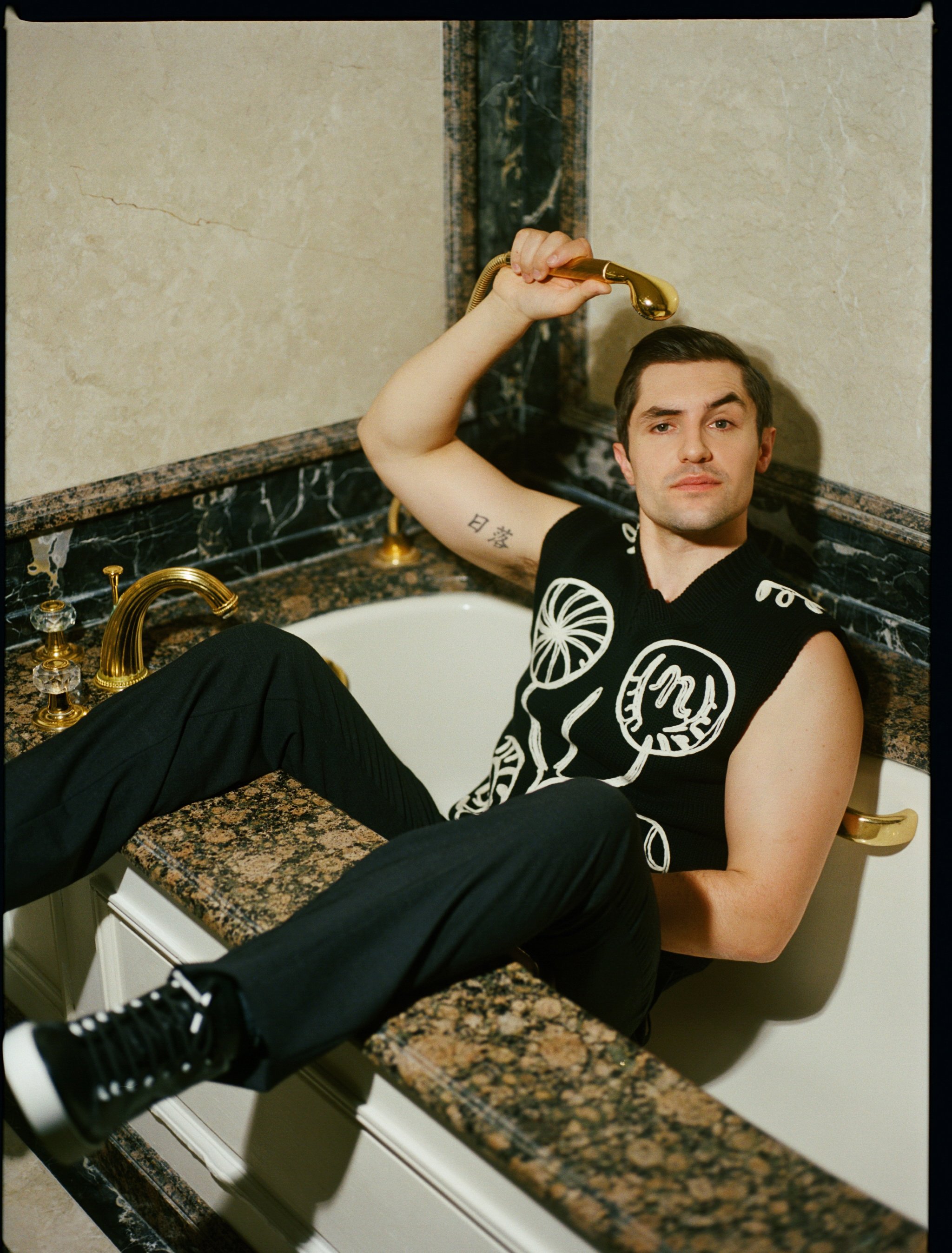 Adult male sits on the edge of a marble bathtub wearing a black tank top and black pants while holding the gold shower head.