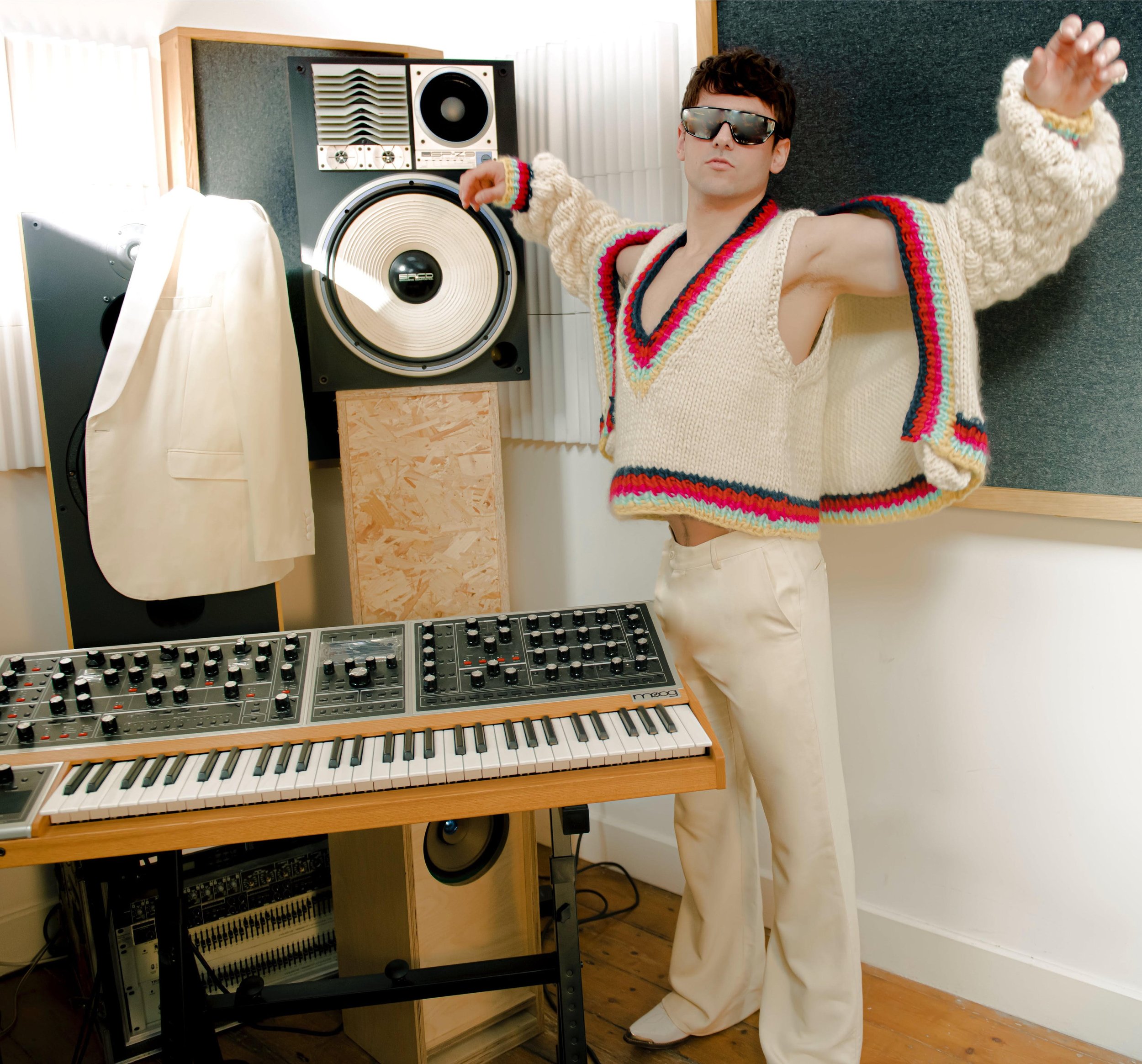 Man in knit sweater vest and trousers stands in front of a keyboard and speakers in a studio, wearing sunglasses and arms spread.