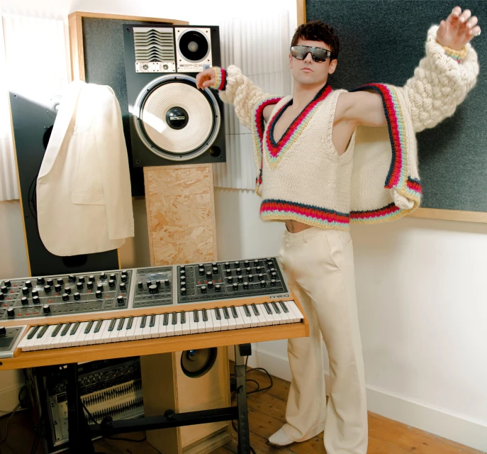 Man in knit sweater vest and trousers stands in front of a keyboard and speakers in a studio, wearing sunglasses and arms spread.