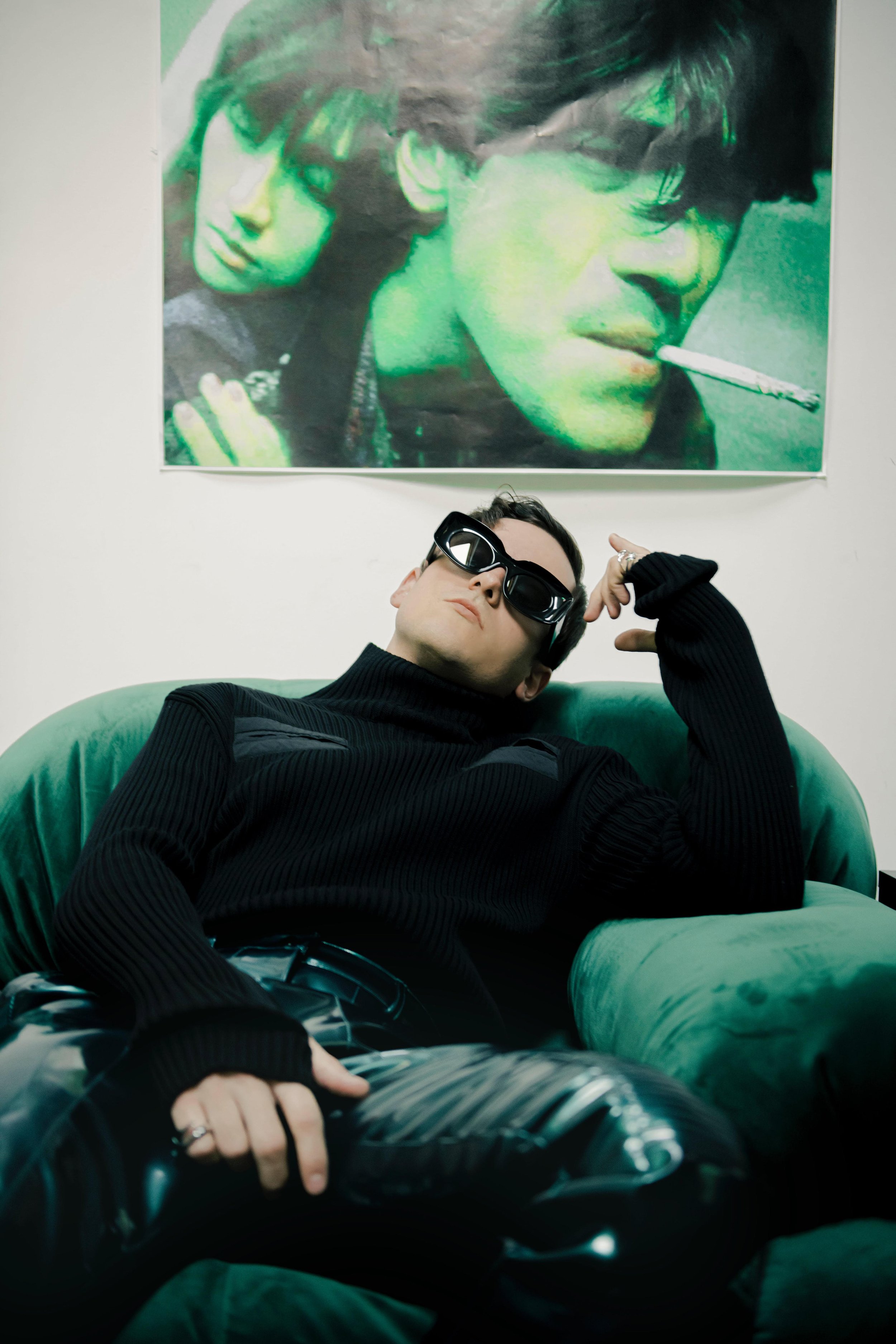 Stylish man in black turtleneck and sunglasses lounges on a green velvet sofa under a portrait. Fashion-forward and contemporary.