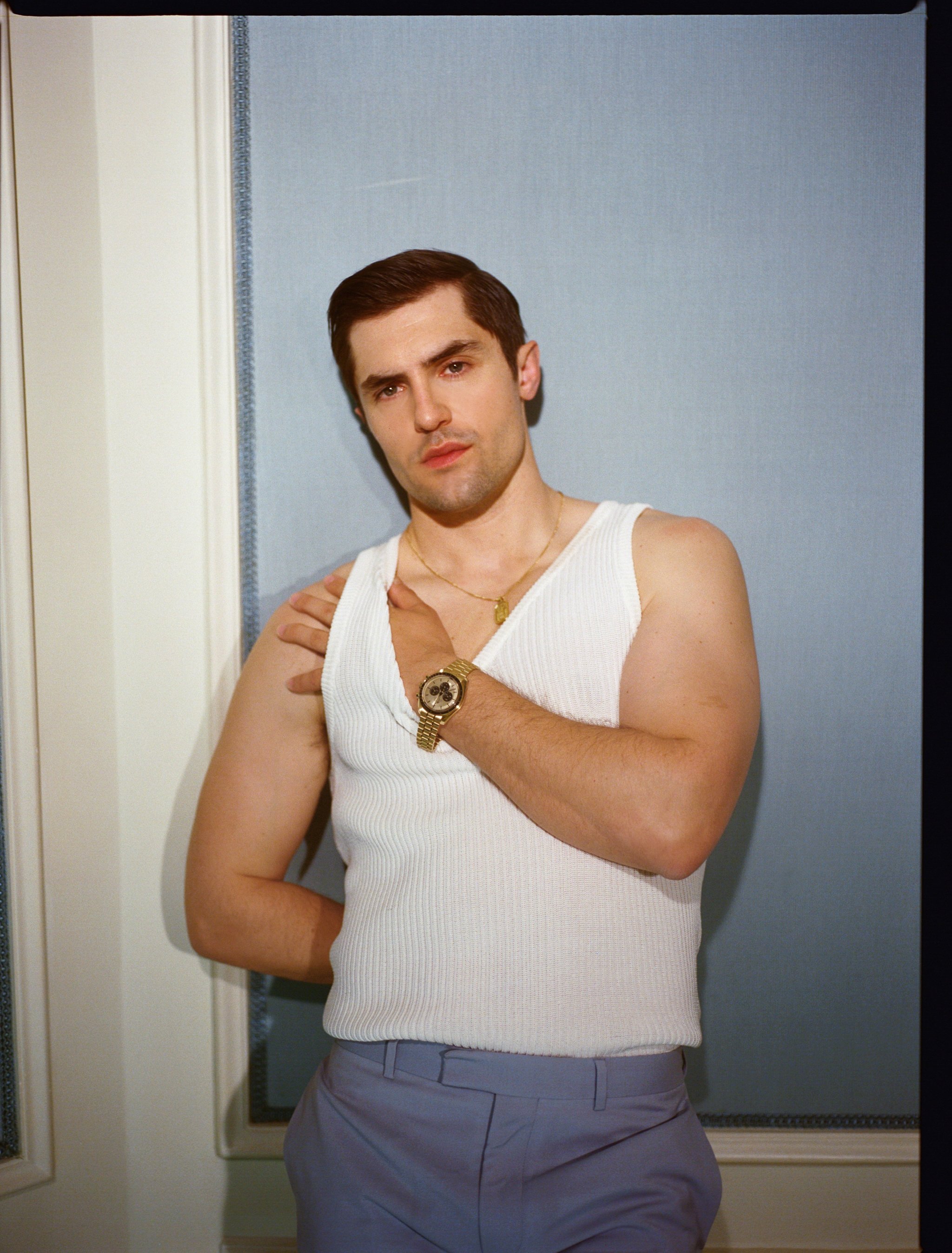 Young adult man in a white tank top and blue trousers poses indoors. He is wearing jewelry with a neutral facial expression.