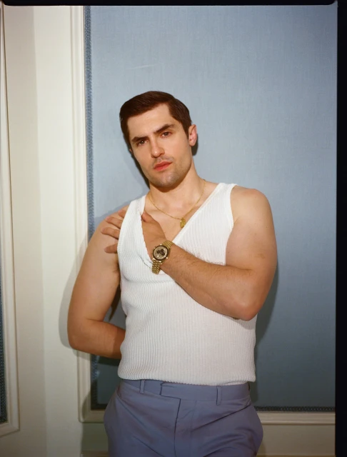 Young adult man in a white tank top and blue trousers poses indoors. He is wearing jewelry with a neutral facial expression.
