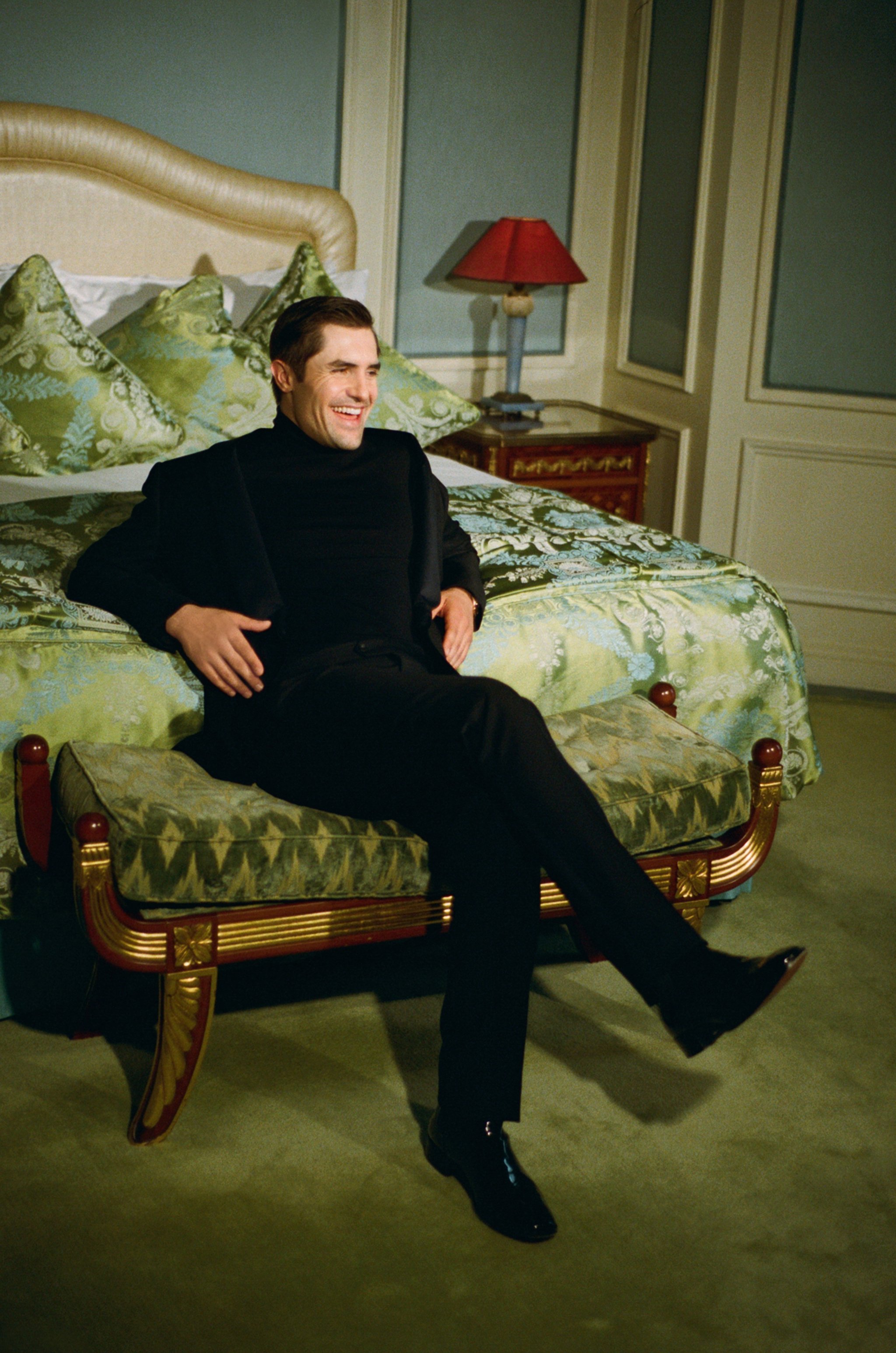 A well-dressed, smiling adult man in a black suit is sitting on a bench in a bedroom with a patterned bed and a light blue wall.
