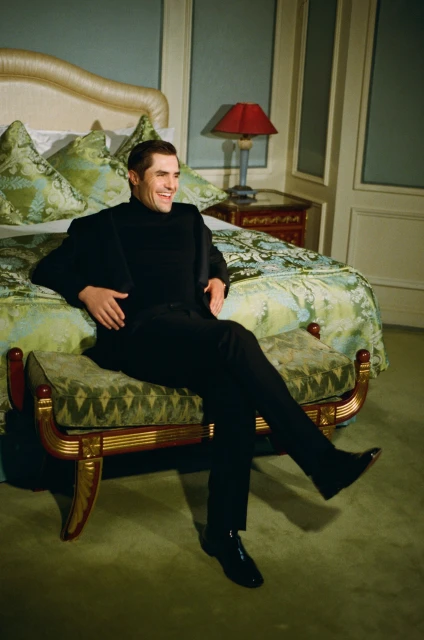 A well-dressed, smiling adult man in a black suit is sitting on a bench in a bedroom with a patterned bed and a light blue wall.
