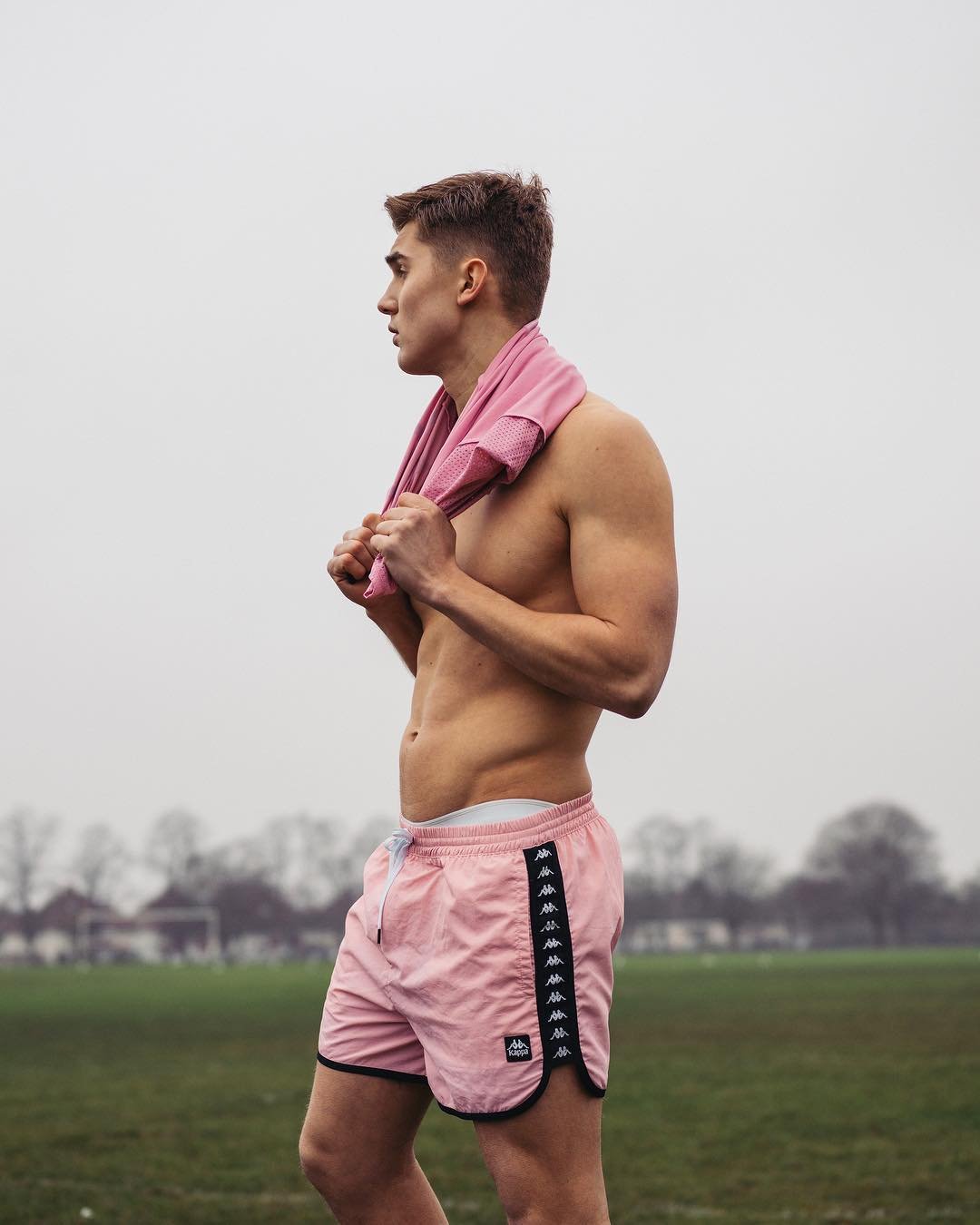 Athletic man outdoors in pink Kappa shorts with a pink towel around his neck, looking toward the left in a lifestyle shot.