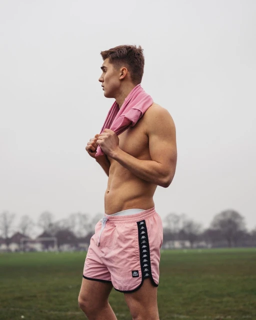 Athletic man outdoors in pink Kappa shorts with a pink towel around his neck, looking toward the left in a lifestyle shot.