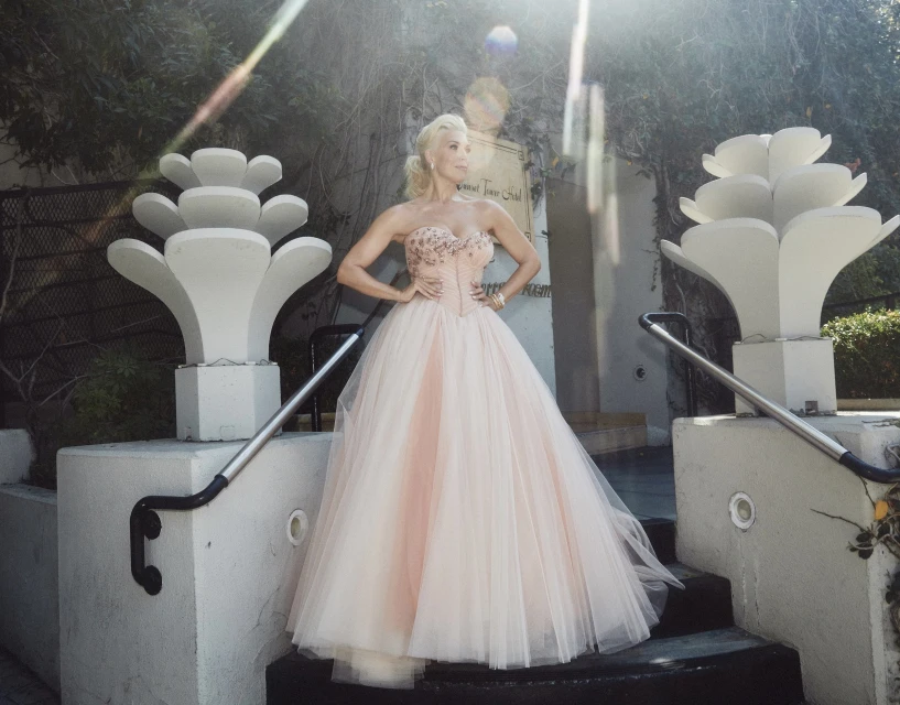 Adult woman in a light pink gown poses on a staircase outdoors, with lens flare, decorative planters, and greenery in the background.