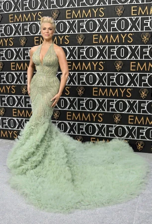Blonde adult female attends an event in a light green halter gown with a ruffled train, posing on a silver carpet in front of a logo wall.