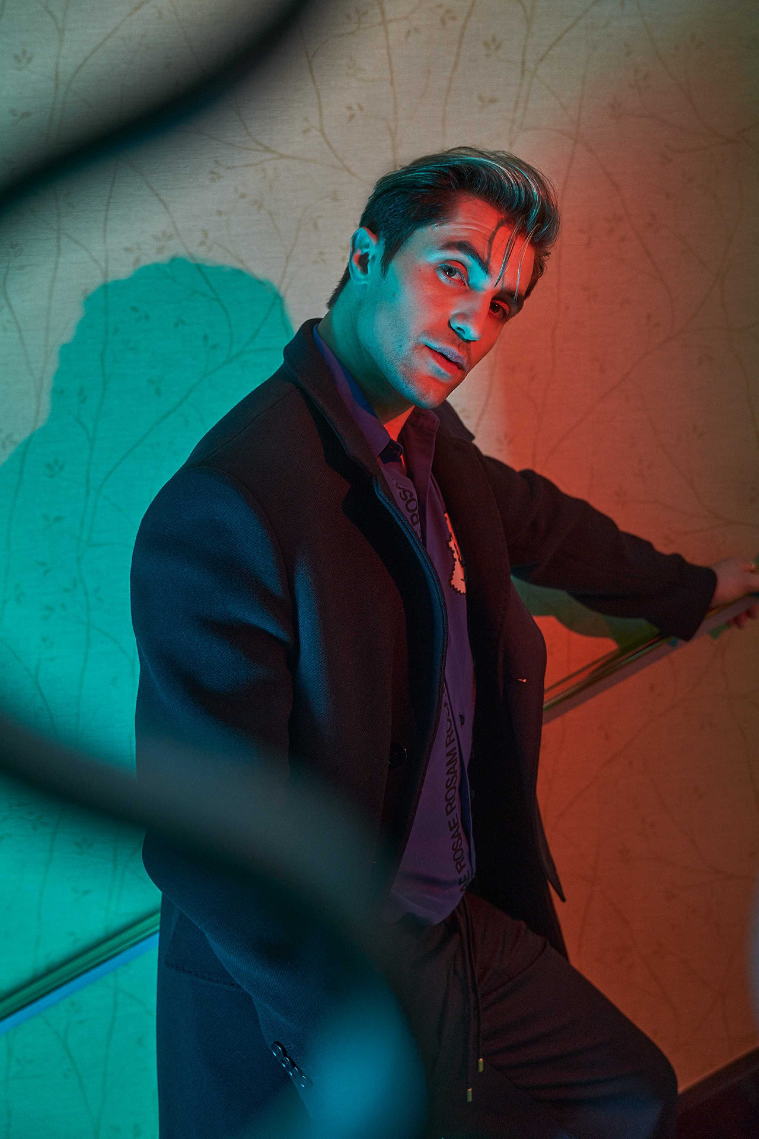 Man in a dark coat and purple shirt posing under dramatic red and green lighting against a patterned wall
