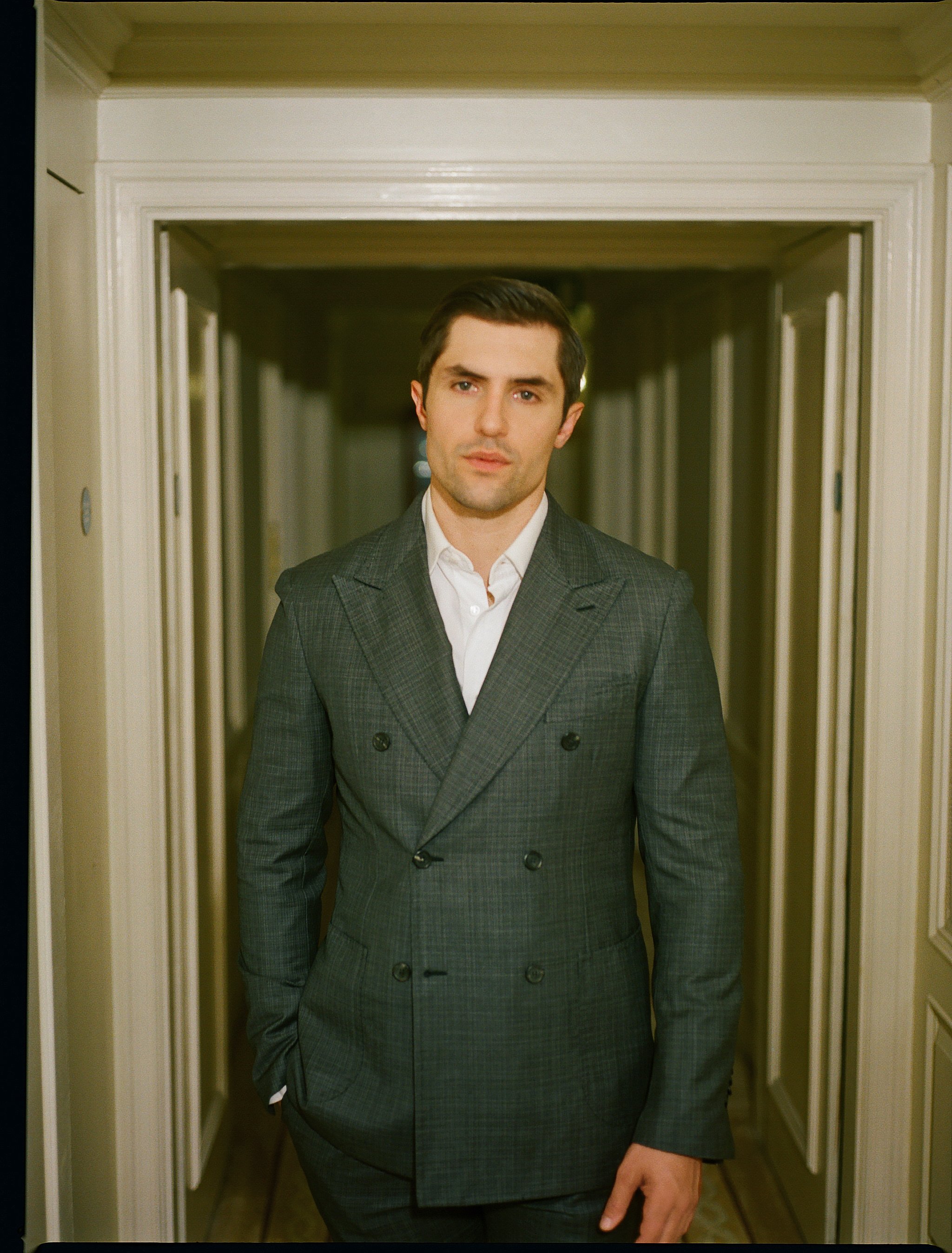 Adult male in a double-breasted suit and white shirt, posing in a hallway with white walls and door frames, looking at the camera.