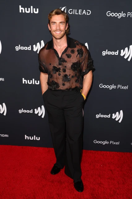 A fair-skinned adult male poses on a red carpet wearing a black, sheer floral button-up shirt with black pants and shoes at an event.