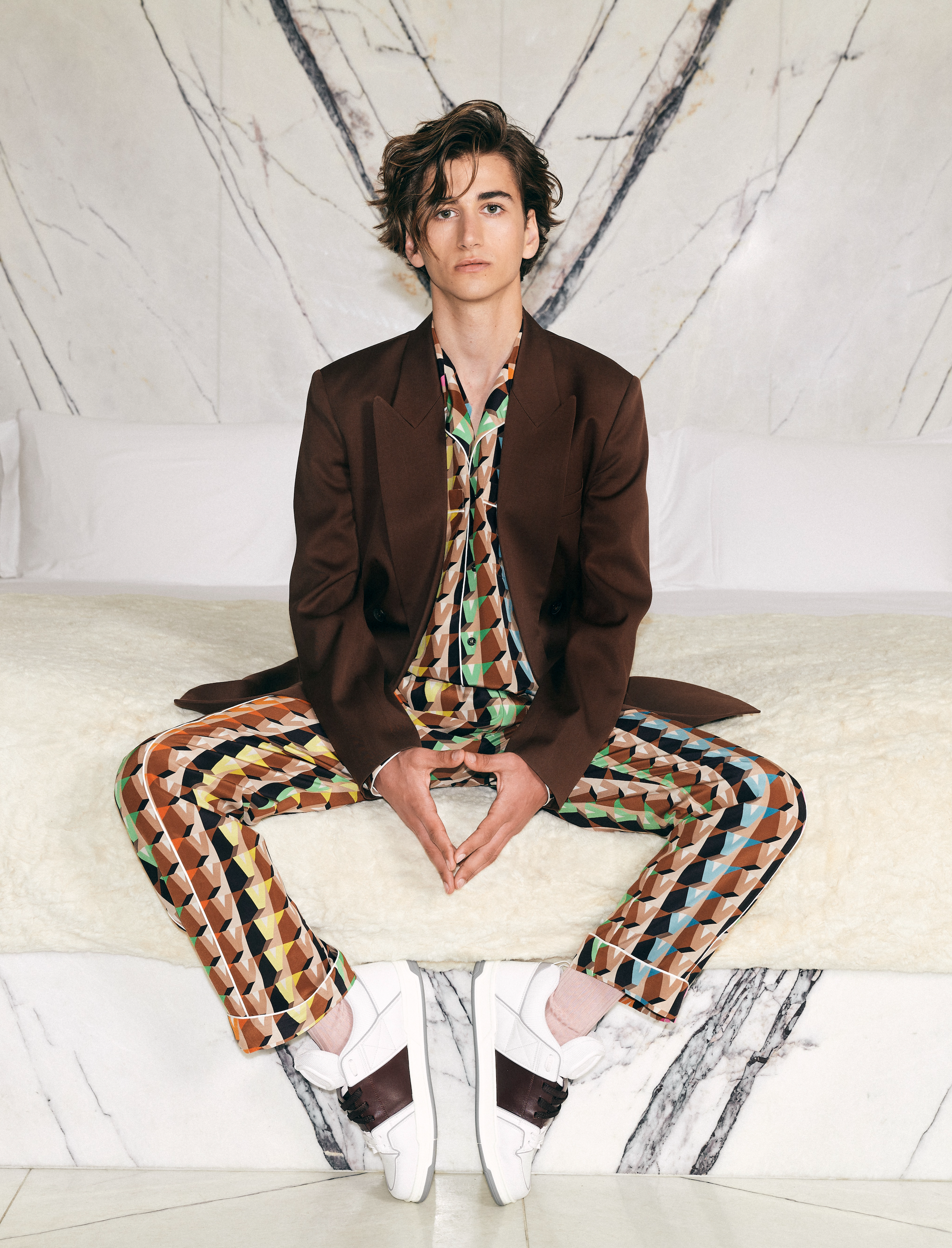 Man in a brown suit and patterned pants sits on a white bed, hands together, white sneakers, with a marble patterned wall in the background.