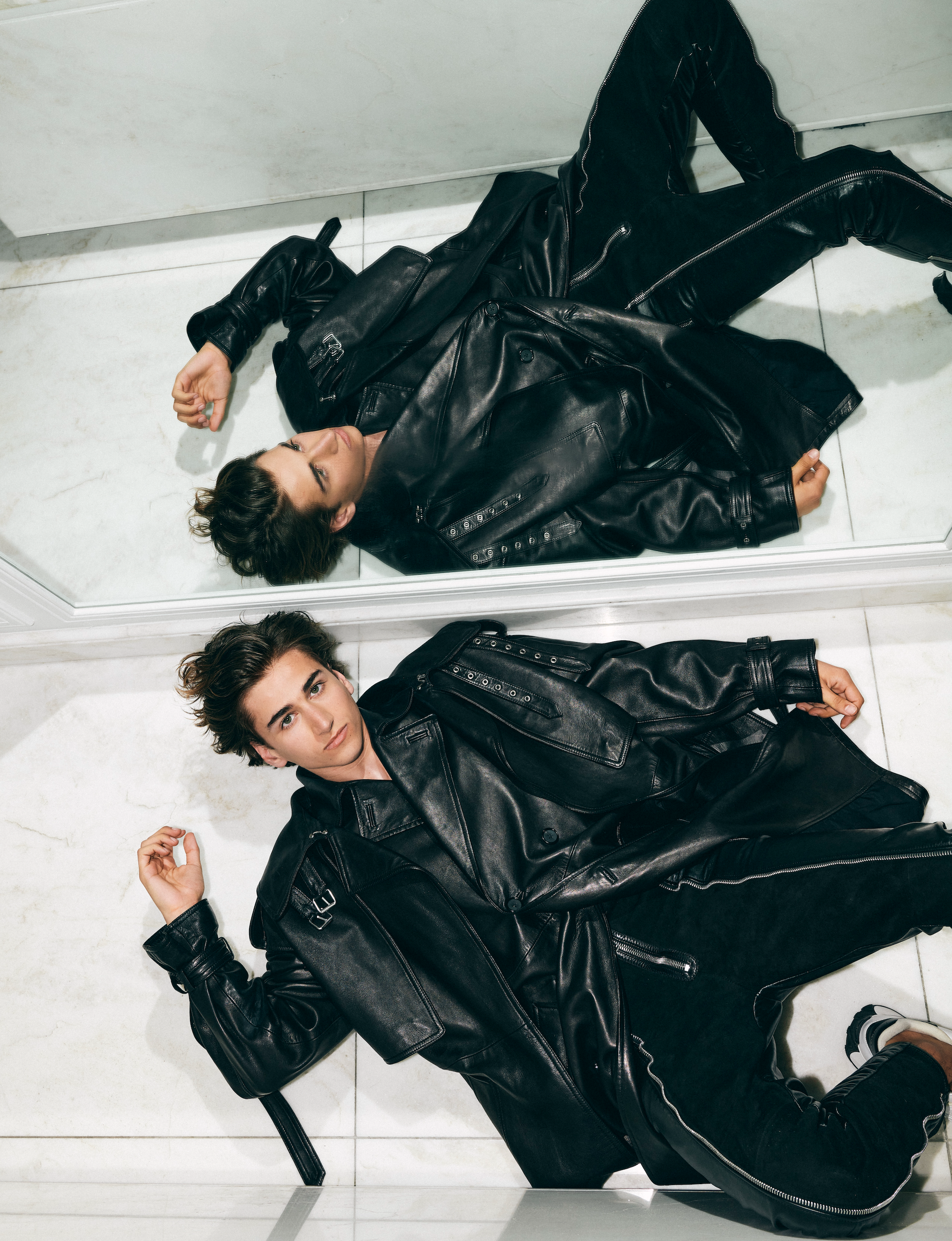 Fashion shot of a young man in a black leather jacket and pants lying on a white tiled floor with his reflection visible above.