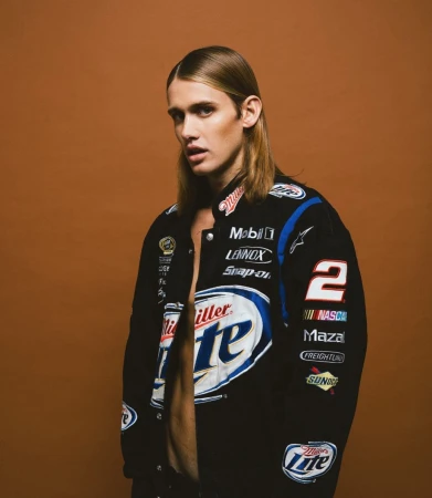Young adult with long hair wearing an unbuttoned racing jacket against a solid brown background in a studio environment.