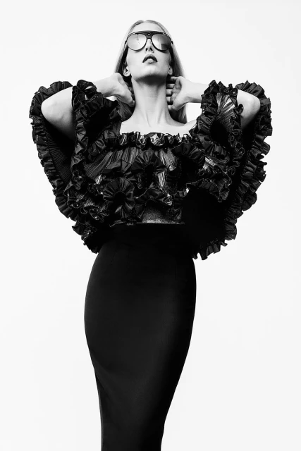 A blonde fashion model in black dress and sunglasses poses with hands behind head against a white backdrop in a studio setting.