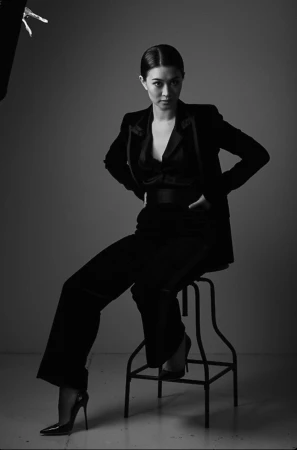 A woman in black business attire sits on a stool in a photo studio, looking directly at the camera in a black and white image.