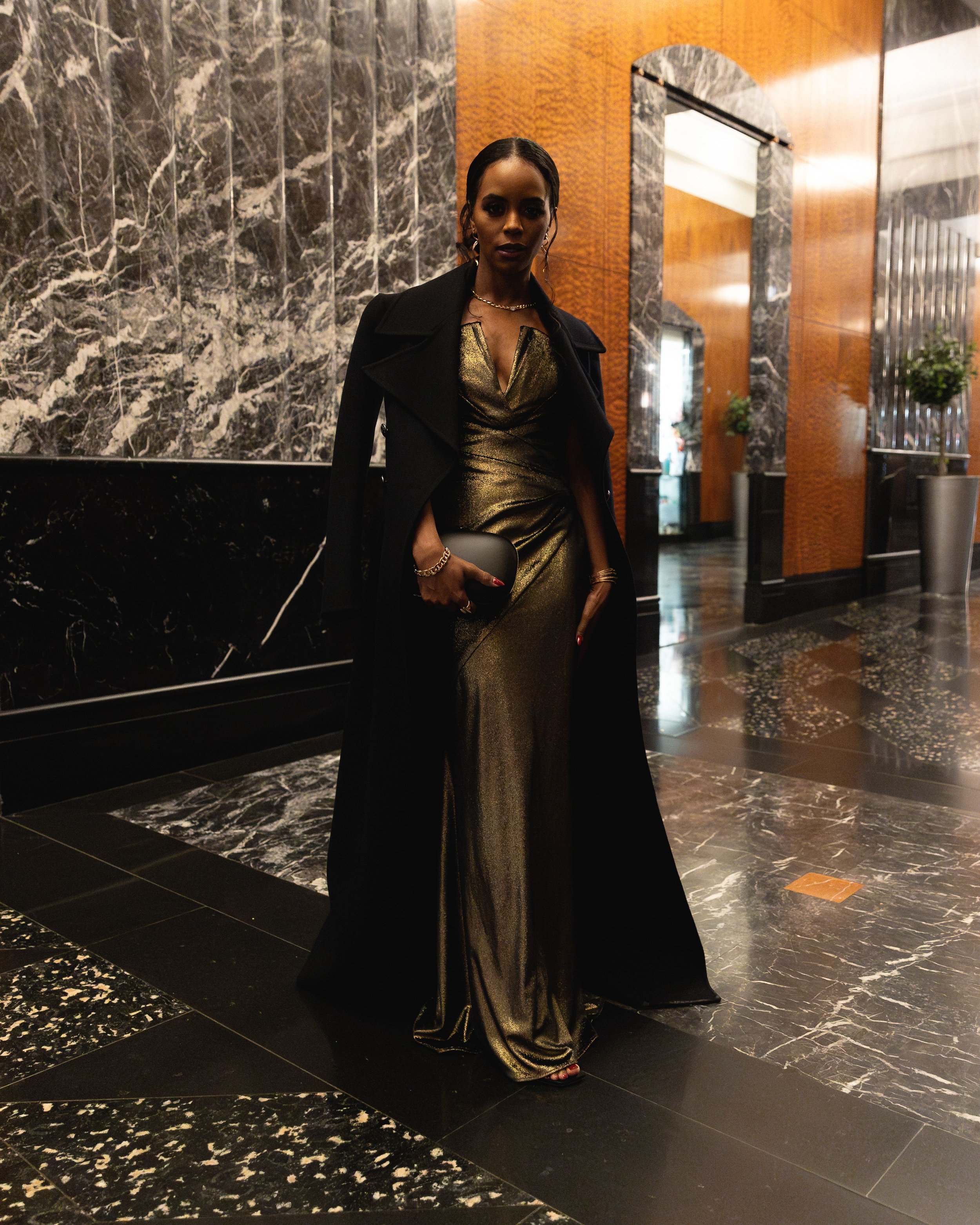 Elegant Black woman in gold gown and black coat poses in a marble-clad foyer, holding a clutch, exuding sophistication and style.