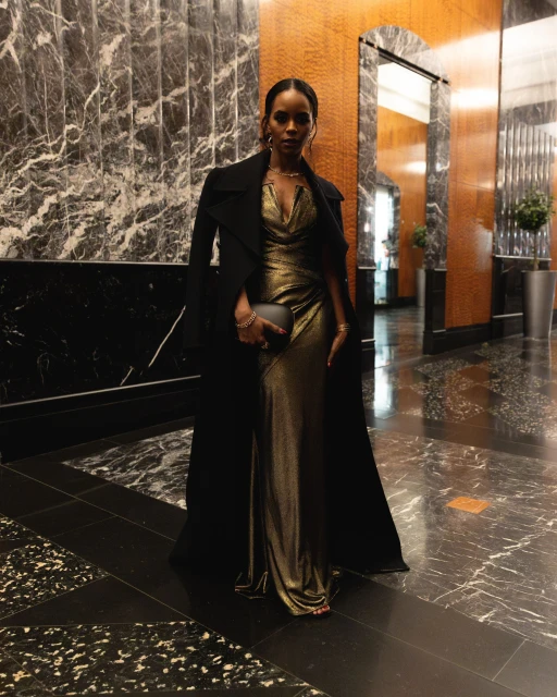 Elegant Black woman in gold gown and black coat poses in a marble-clad foyer, holding a clutch, exuding sophistication and style.