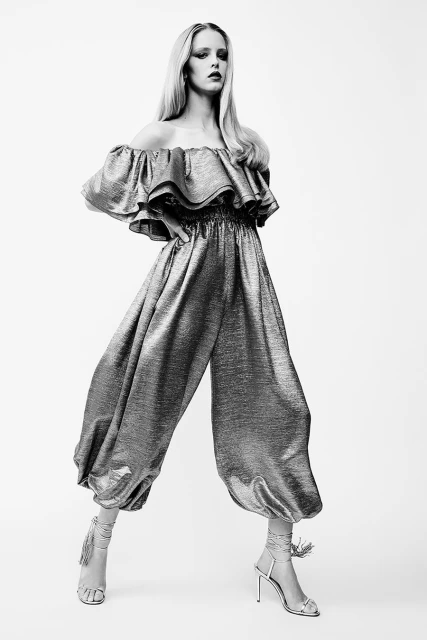 Elegant blonde woman in shimmering metallic off-the-shoulder jumpsuit and stiletto heels, posing against a bright white backdrop in a studio.