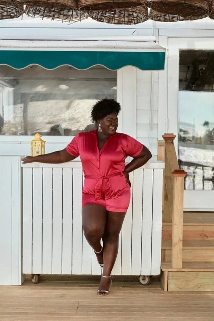 Attractive adult female in a fashionable pink romper with matching heels posing in front of a white building on a sunny day.